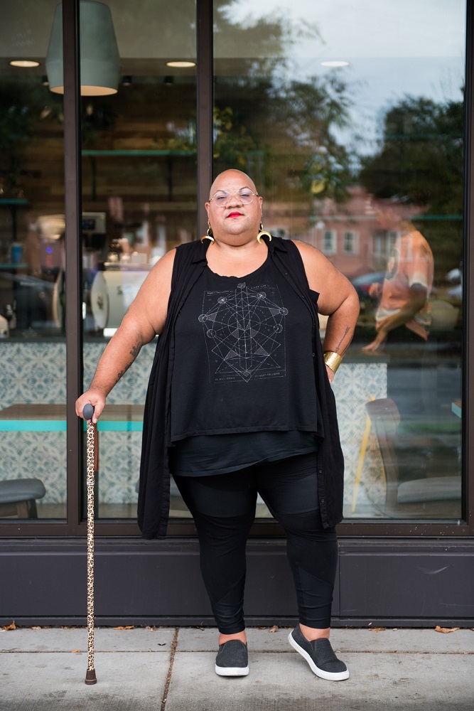 A Black non-binary person stands casually outside a cafe while leaning on their leopard print cane. They are dressed in all black and have a shaved head, glasses, and a red lip, along with moon earrings and a gold bracelet.