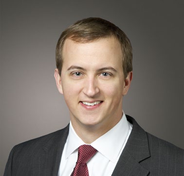 A white man in a suit and tie smiles in front of a gray background.