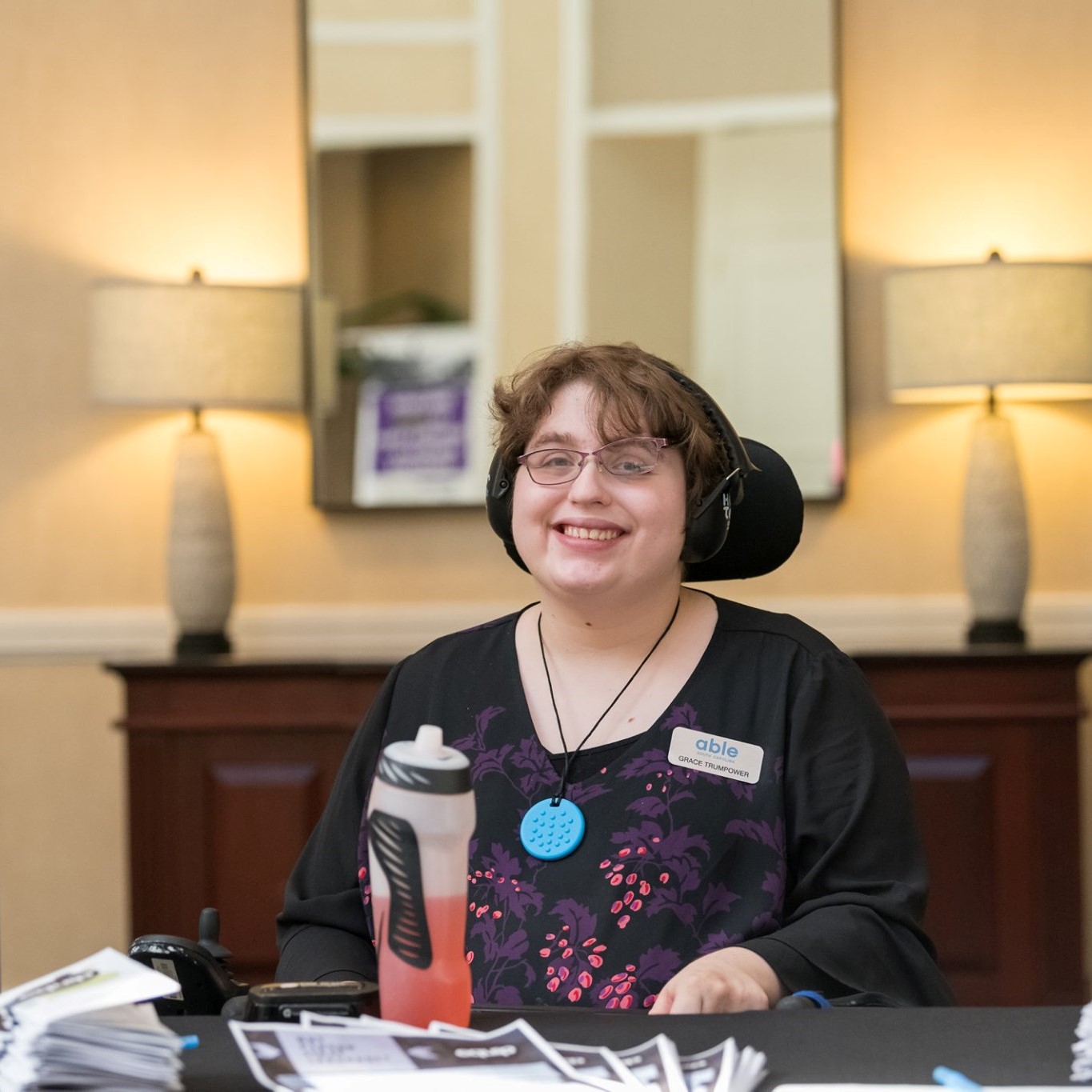 Grace wears glasses, headphones, and a black top while smiling and sitting in a wheelchair behind a vendor table.