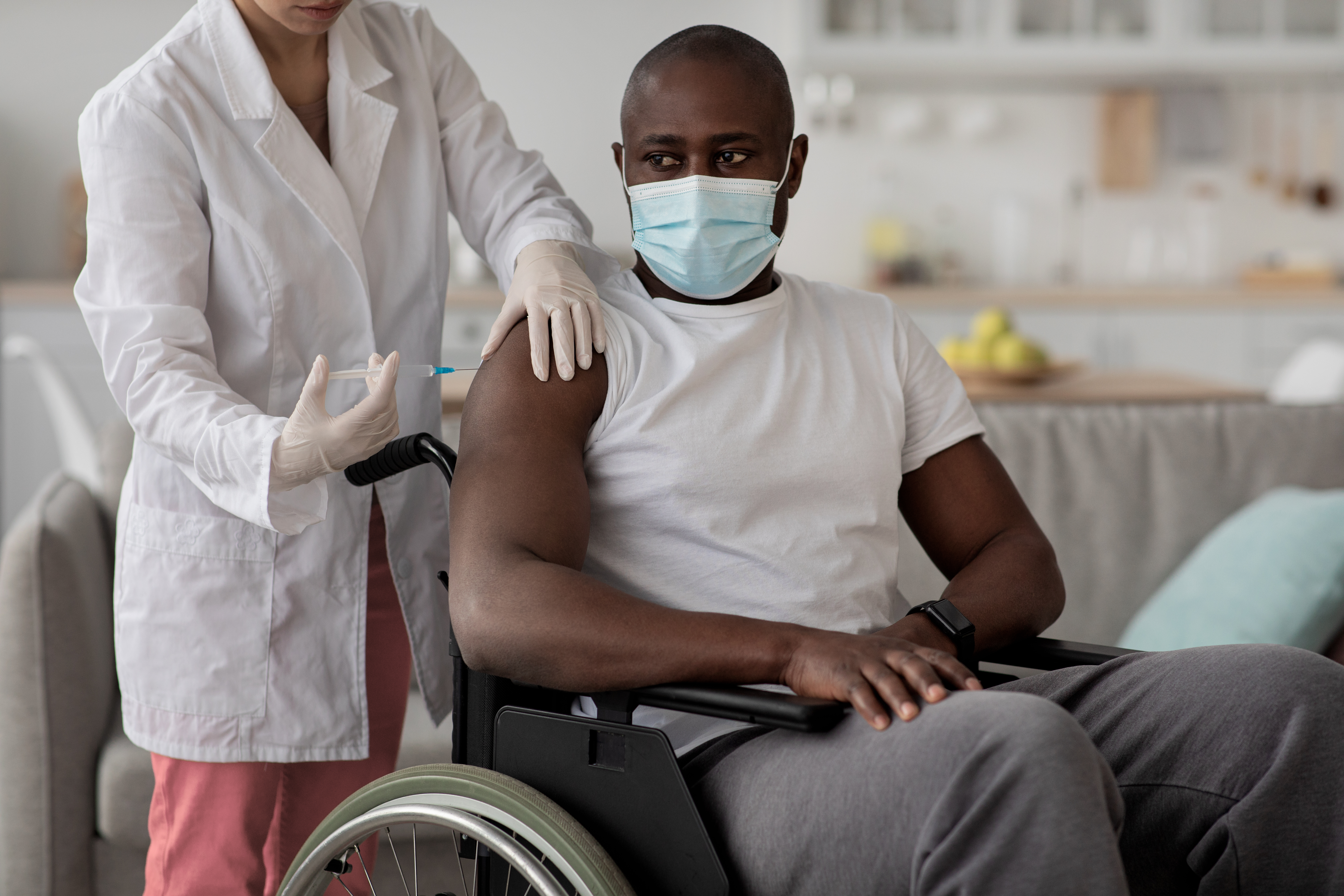 Home health care, community vaccination, influenza immunization and covid-19 pandemic. Paramedic in rubber gloves gives injection to black man in protective mask in wheelchair in living room interior