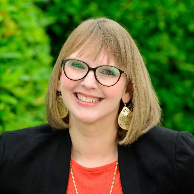 Kimberly is a white woman with shoulder lenght blonde hair with bangs. She is wearing brown rounded rim glasses, gold teardrop earrings, and gold necklace with a read shirt and black blazer. She smiles for the camera in front of greenery.