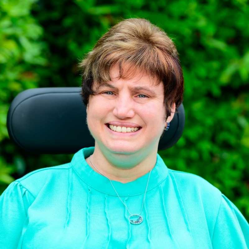 Dori is a white woman with cropped brown hair with blond highlights. She is wearing silver hoop earrings with blue crystals and a matching silver necklace with silver hoop pendant and blue, green, and purple crystal beads, and a turquoise bouse. She is smiling at the camera, from her power wheelchair in front of greenery.