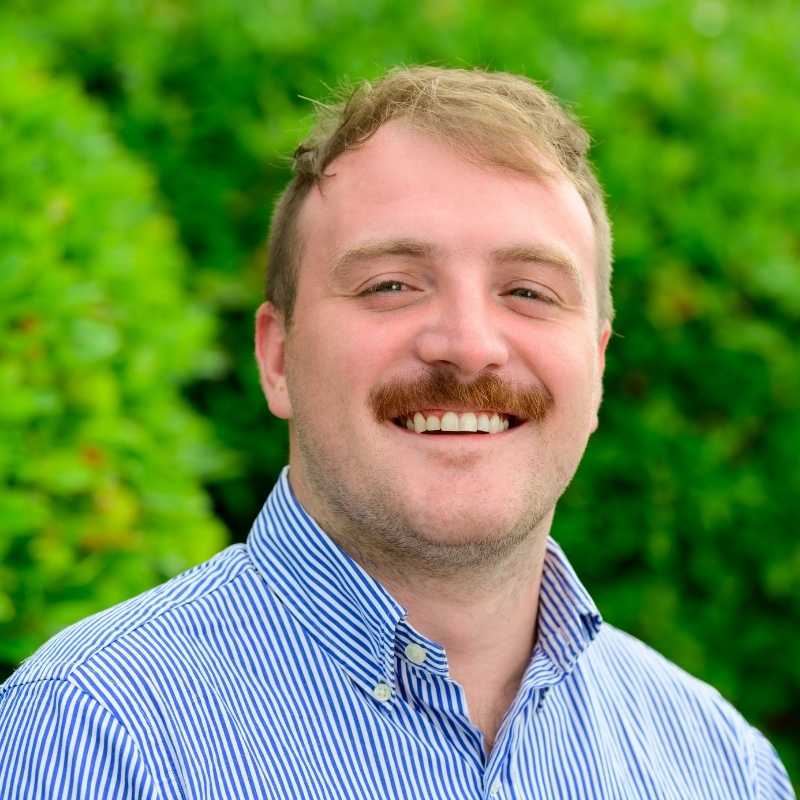 Ryan is a white man with cropped blonde hair and mustache. He is wearing a blue and white striped button-down shirt. He is smiling at the camera, standing in front of greenery.