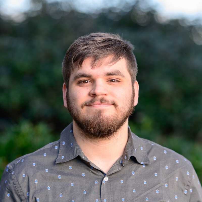 Holden is a white man with brown hair and beard. He is wearing a gray collared shirt with blue dots and smiling in front of greenery outside.