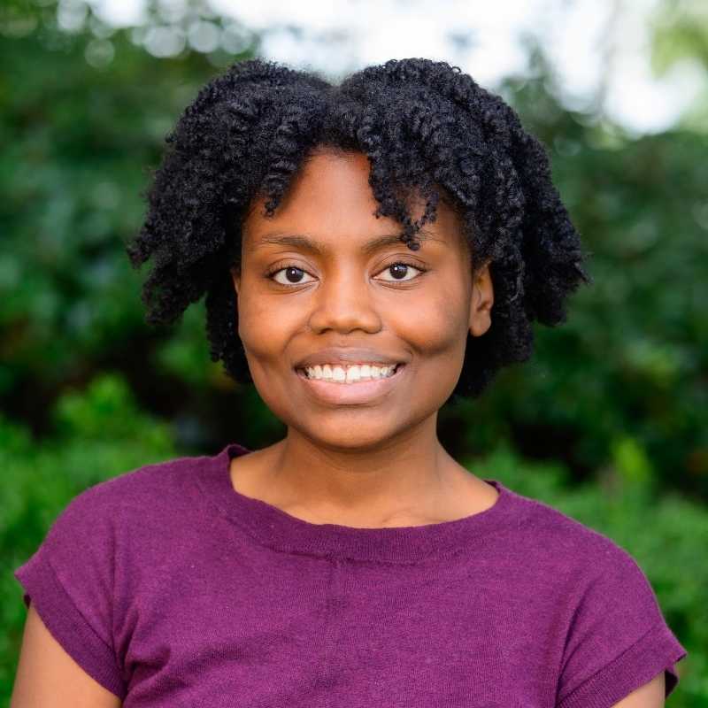 Wednesday is a Black person with black, twisted, kinky natural hair. She is wearing a purple shirt. They are smiling at the camera, standing in front of greenery.