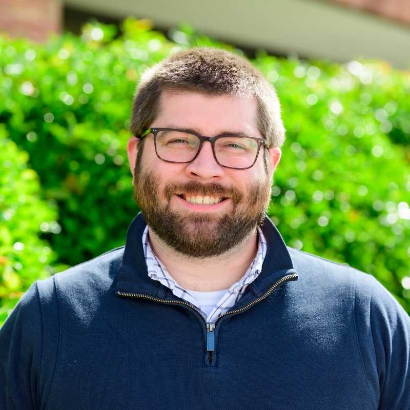 Michael is a white man with brown cropped hair and closely trimmed beard with brown rimmed glasses. He is wearing a navy quarter zip sweater over a white and blue checked collared shirt. He is smiling at the camera, standing in front of greenery.