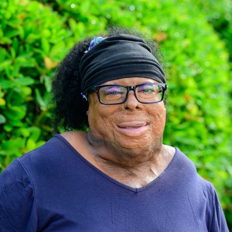 LaQuanda is a Black woman with black kinky hair tied back in a black headband, wearing black rectangular framed glasses that show off her punchy purple eye shadow, and a navy shirt. She is smiling at the camera, standing in front of greenery.