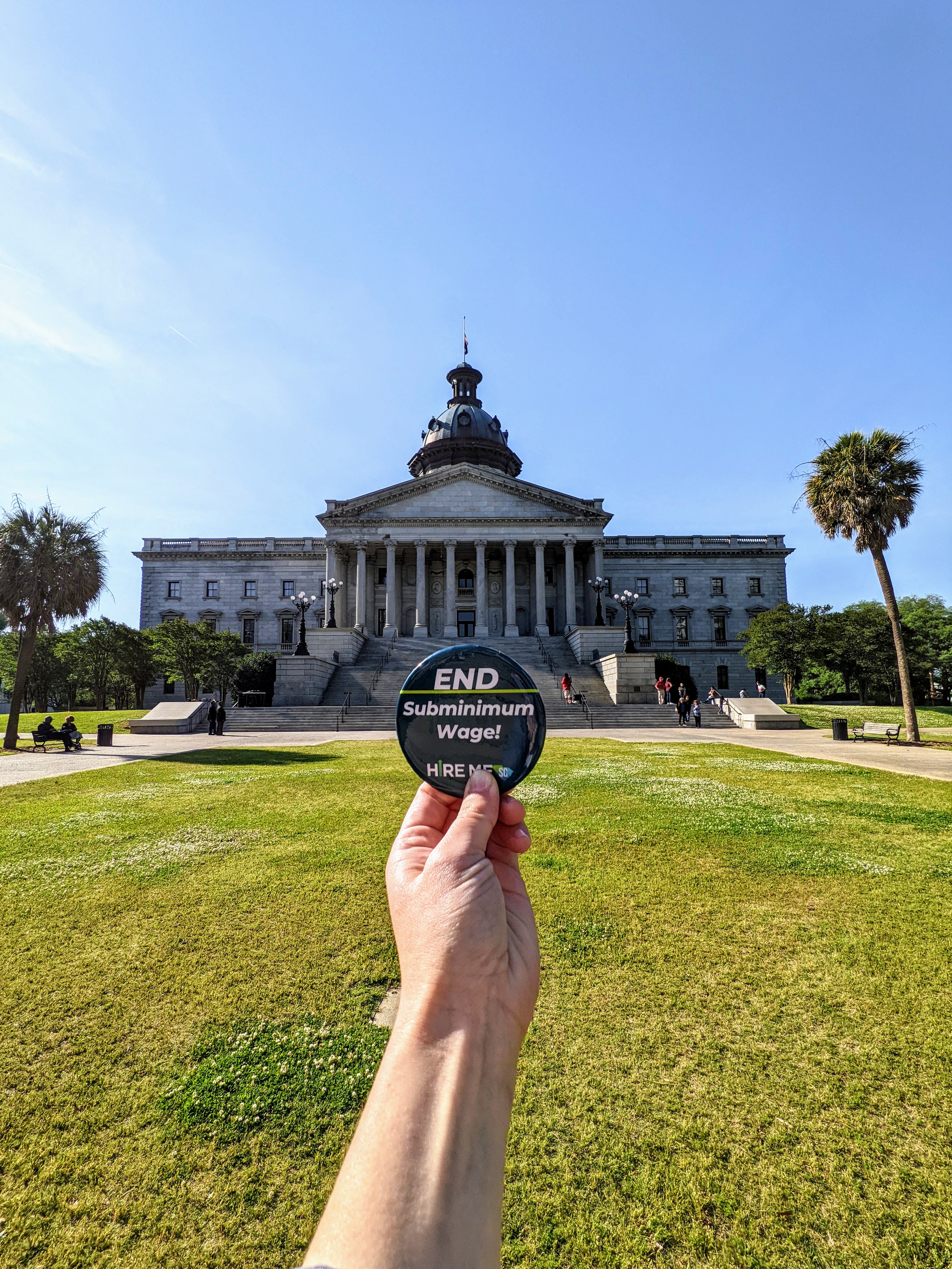 Photograph of South Carolina State House in the background behind a hand holding a button that reads 'End subminimum wage,' with the hire me sc logo, in the foreground.