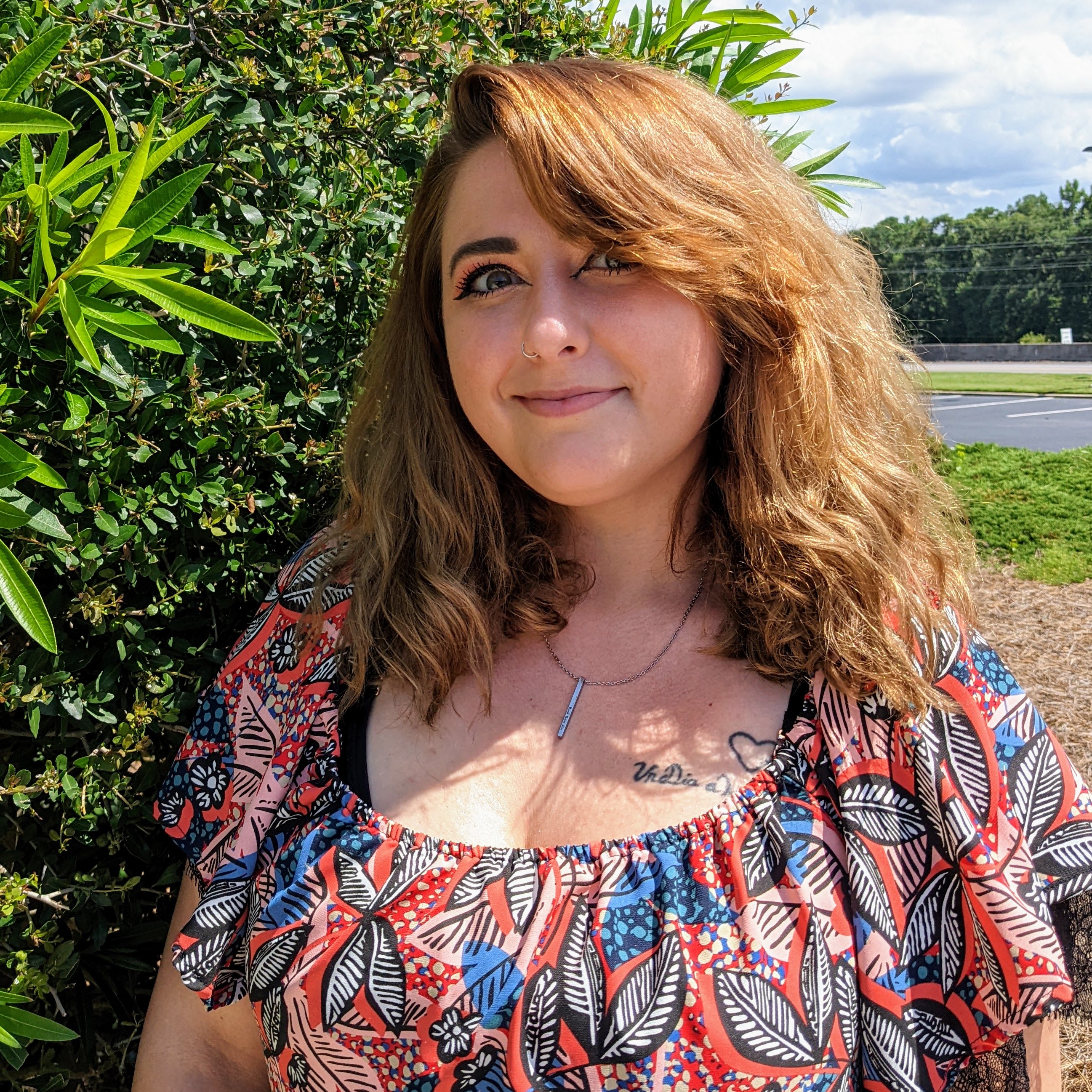 Savvy is a white woman with long brown hair with a red tint. She is wearing a floral blouse and smiling with a closed mouth while standing in front of greenery outside.