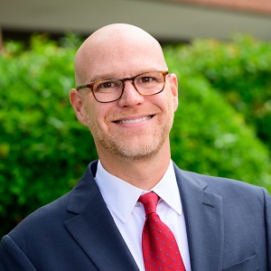 Cory, a white man with no hair, light colored beard, and brown glasses. He is wearing a dark blazer with red tie while smiling outside.