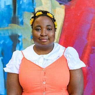 Alexis is a young Black person with cropped natural hair wearing a yellow flower headband with a orange dress with white collared shirt. They are in front of a colorful mural and are smiling closed mouthed.