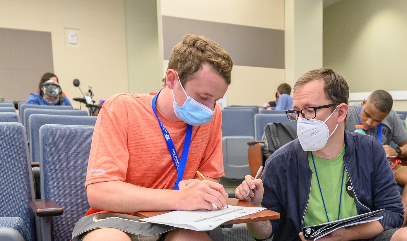Young white student with disabilities works on a worksheet with Able SC staff member, Troy Hall. Troy is a white man with glasses. They are both wearing facemasks and casual clothing in a classroom setting.