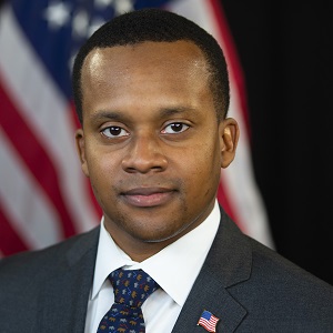 Scott, a young Black man with cropped hair wearing a suit and tie with an American flag pin and flag in the backround. He looks assuredly at the camera.