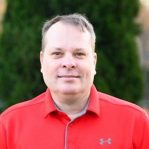 Aaron, a white man with cropped brown hair wearing a red collared shirt and smiling with closed mouth while standing in front of greenery outside.