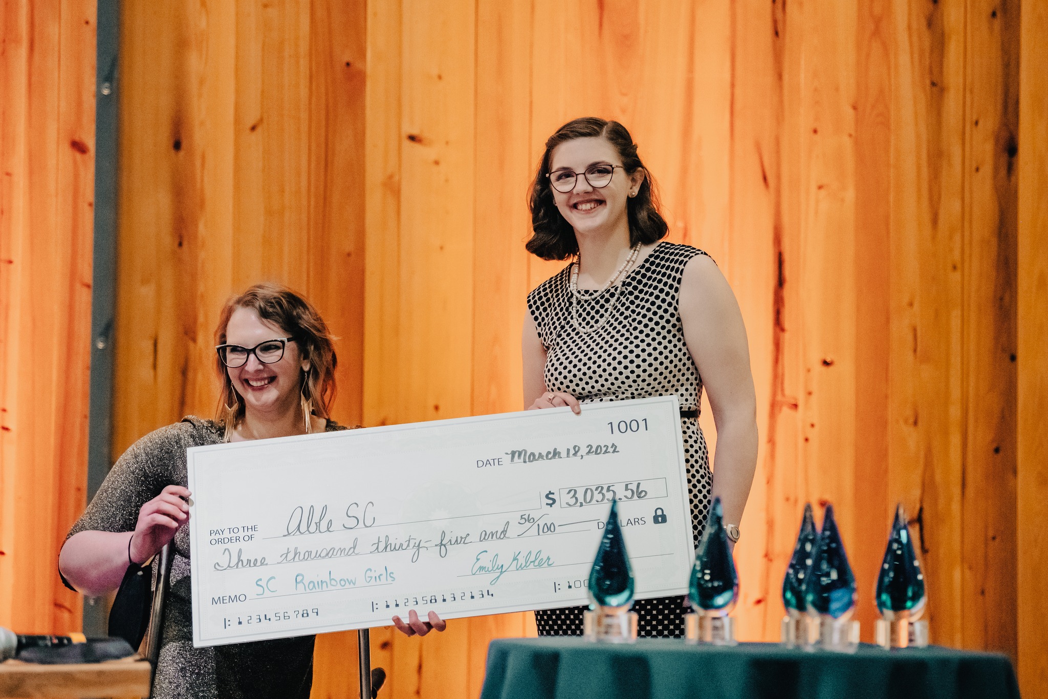 Photo of Kimberly, Able SC's CEO, holding a large check with special guest and fundraiser, Emily. They are standing on the stage and smiling towards the crowd.