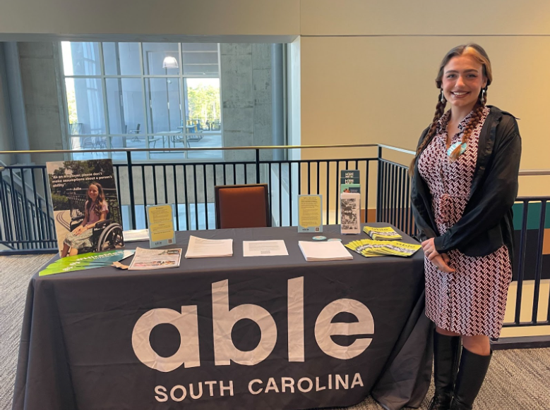 AmeriCorps member standing next to a table with Able SC on the tablecloth and a variety of materials from the organization. She is a white woman with long braided red hair.
