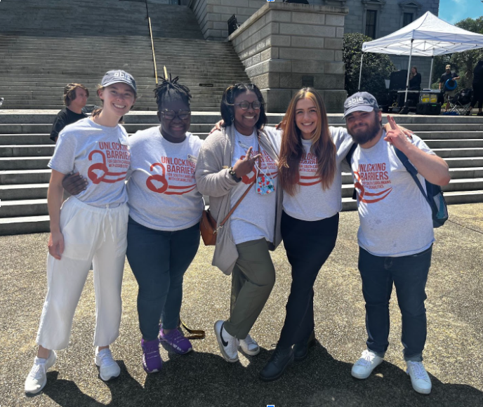 5 AmeriCorps members posing together during Advocacy Day for Access and Independence at the SC Statehouse. From left to right, a white woman with a baseball cap, a Black woman with glasses, a Black person with glasses making a peace sign, a White woman with long red hair, a white man with a beard and ball cap. All are wearing the same official event t-shirt. Shirt is gray with slogan, 'Unlocking barriers for South Carolinians with disabilities,' followed by an unlocked pad lock with the state of SC as the keyhole followed by a swoosh. Print is in burn orange color.
