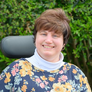 Dori, a white woman in a power wheelchair with cropped brown hair wearing a floral blouse, smiling in front of greenery outside.
