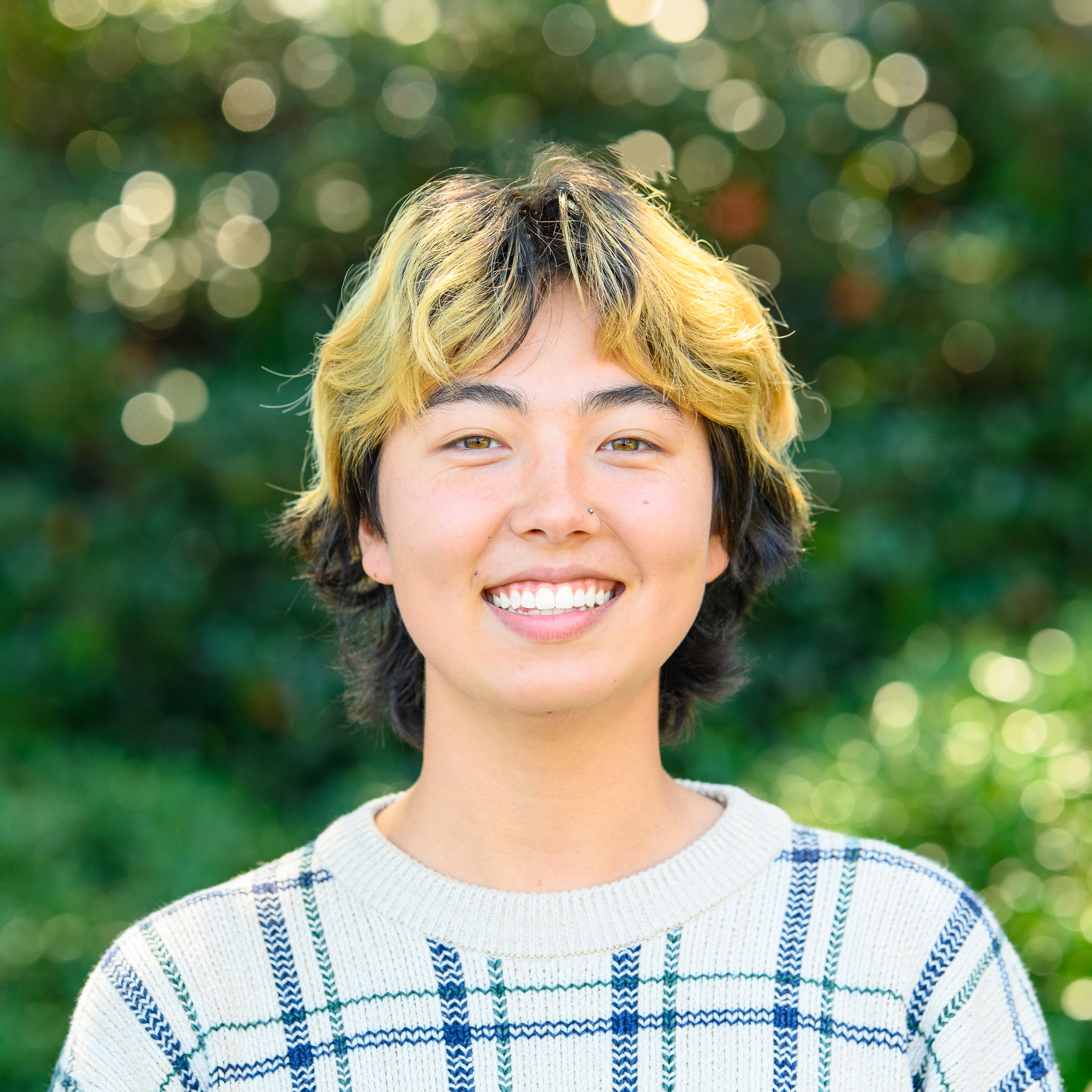 Photo of Sun, a Korean-American person with cropped black hair with gold highlights. They are smiling outside in front of greenery.