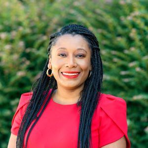 McKenzie is a Black woman with long black braids. She is wearing hoop earrings and a red top with matching red lip color, while smiling in front of greenery outside.