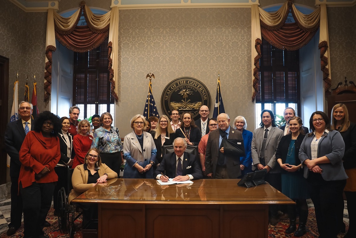 Able SC staff, consumers, supporting partner organizations, lawmakers, and advocates pose smiling for a photo behind the Governor's desk as he signs a bill to end subminimum wage and support Employment First. The people pictured are diverse in age, race, gender, and disability.