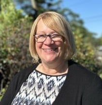 Sue, a white woman with blonde hair and glasses smiling while outside.