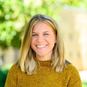 Aimee, a woman with blonde hair and a gold sweater, smiles while standing outside.
