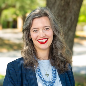Taylor, a white woman with brown-gray hair in a blue suit wearing red lipstick, smiling outside.