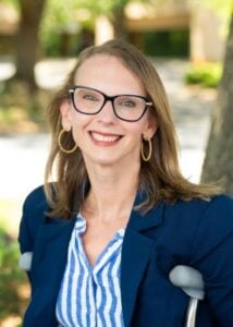 Kimberly, a white woman with long brown hair and glasses smiling outside. The crutches she uses to walk are visible.