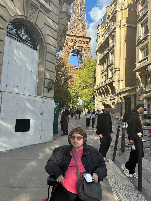  a photo of the author, a white person with short brown hair, glasses, and headphones, sitting in their wheelchair on a sidewalk in Paris. They are in between two buildings, and many people are standing around behind them in the background. The Eiffel Tower is also visible in the background.
