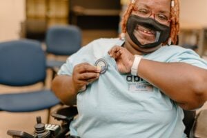 Black woman in an Able SC shirt wearing a facemask with a clear view center puts on a button promoting their wellness through vaccination.