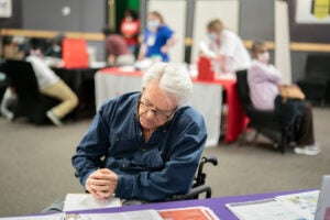White man with gray hair in a wheelchair fills out a form at a clinic.