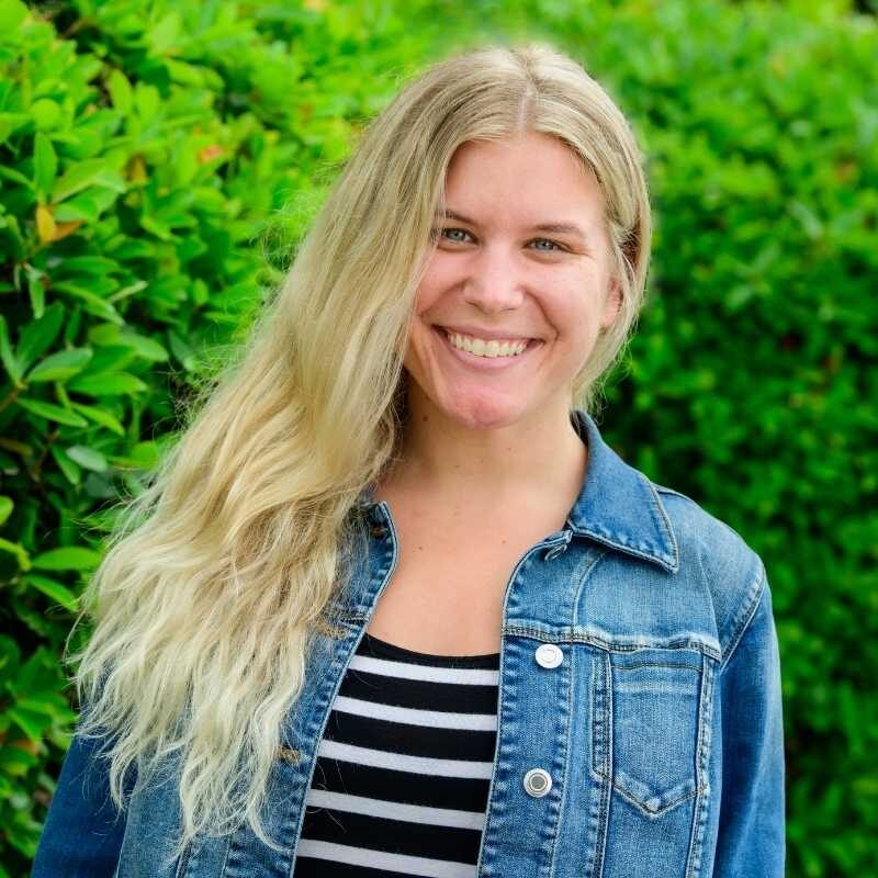 Aimee is a white woman with long wavy light blonde hair. She is wearing black and white striped shirt, layered with a blue jean jacket. She is smiling at the camera, standing in front of greenery.