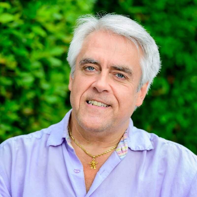 Mark is a white man with short white hair. He is wearing a gold necklace with frog pendant and lavender collared shirt. He is smiling at the camera, in front of greenery.