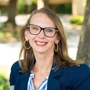 Kimberly, a white woman with long brown-blonde hair and glasses smiling while outside- she is wearing a blue blazer and hoop earrings.