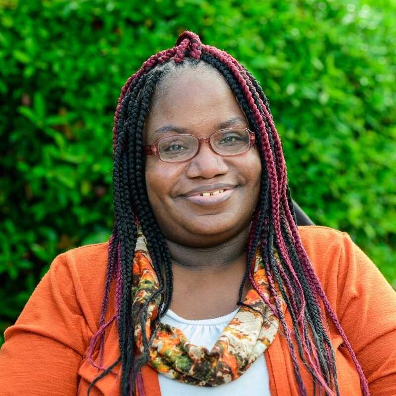 Hope is a Black woman with black and burgundy long braids with translucent brown rimmed glasses. She is wearing an orange cardigan layered over a white shirt with an orange, cream, yellow, and green abstract printed scarf. She is smiling at the camera from her power wheelchair in front of greenery.