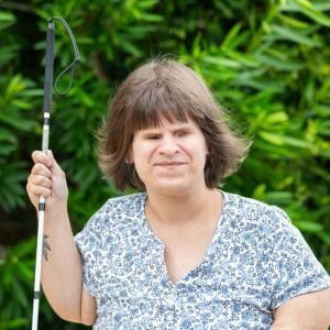 Ashley, a blind woman with short brown hair and a flower top holding a long white cane outside.