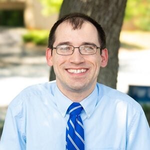 Tyler, a white man with glasses and brown cropped hair in a blue shirt and tie, smiling outside.