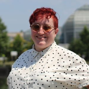 Abby, a white person with cropped red dyed hair wearing sunglasses and a white blouse with black polka dots. They smile while standing outside.