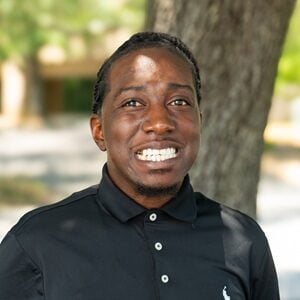 Donovann, a Black man with long dreds pulled back, smiling in a dark shirt while outside.