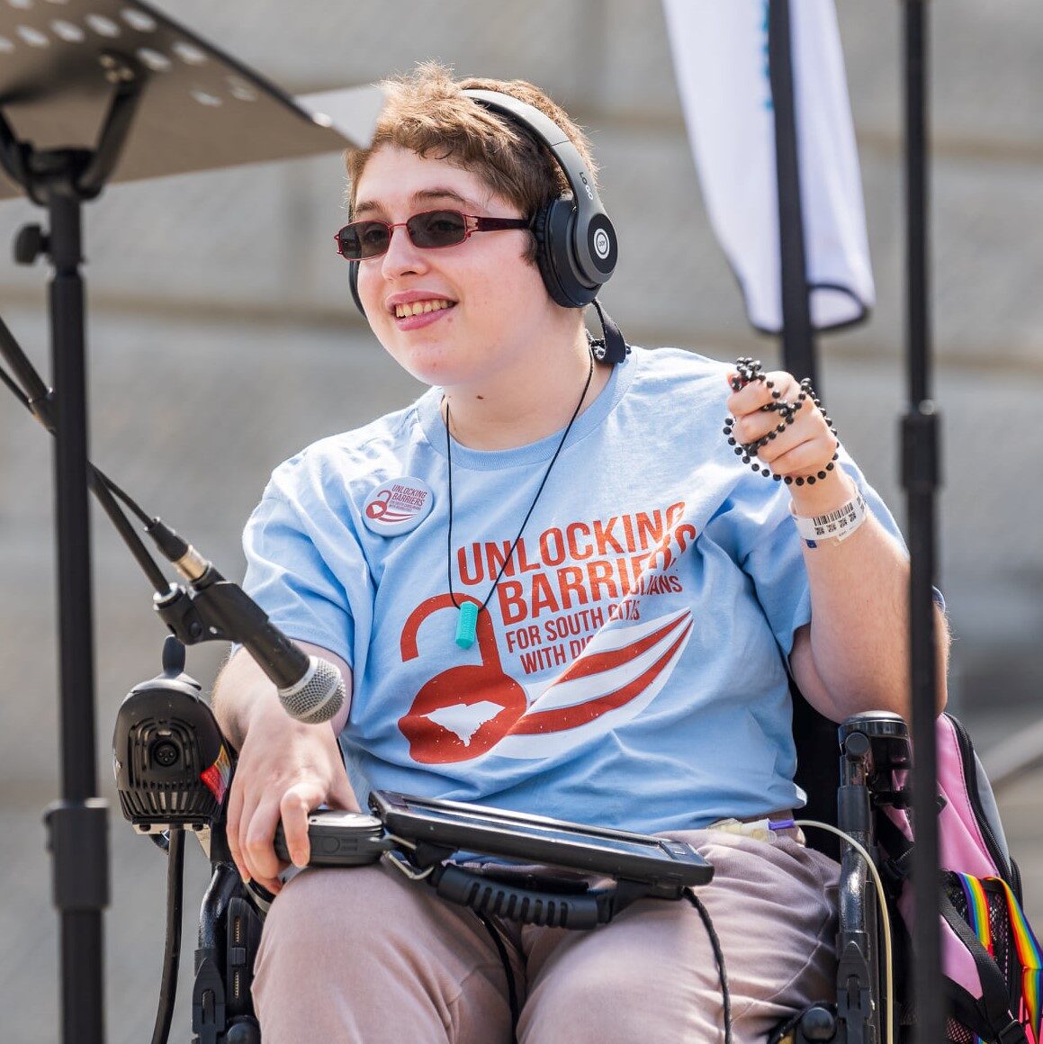 Grace is a young white person with cropped brown hair and sunglasses wearing headphones and using assistive speech techonology in their power wheelchair while giving a speech on the SC statehouse steps.